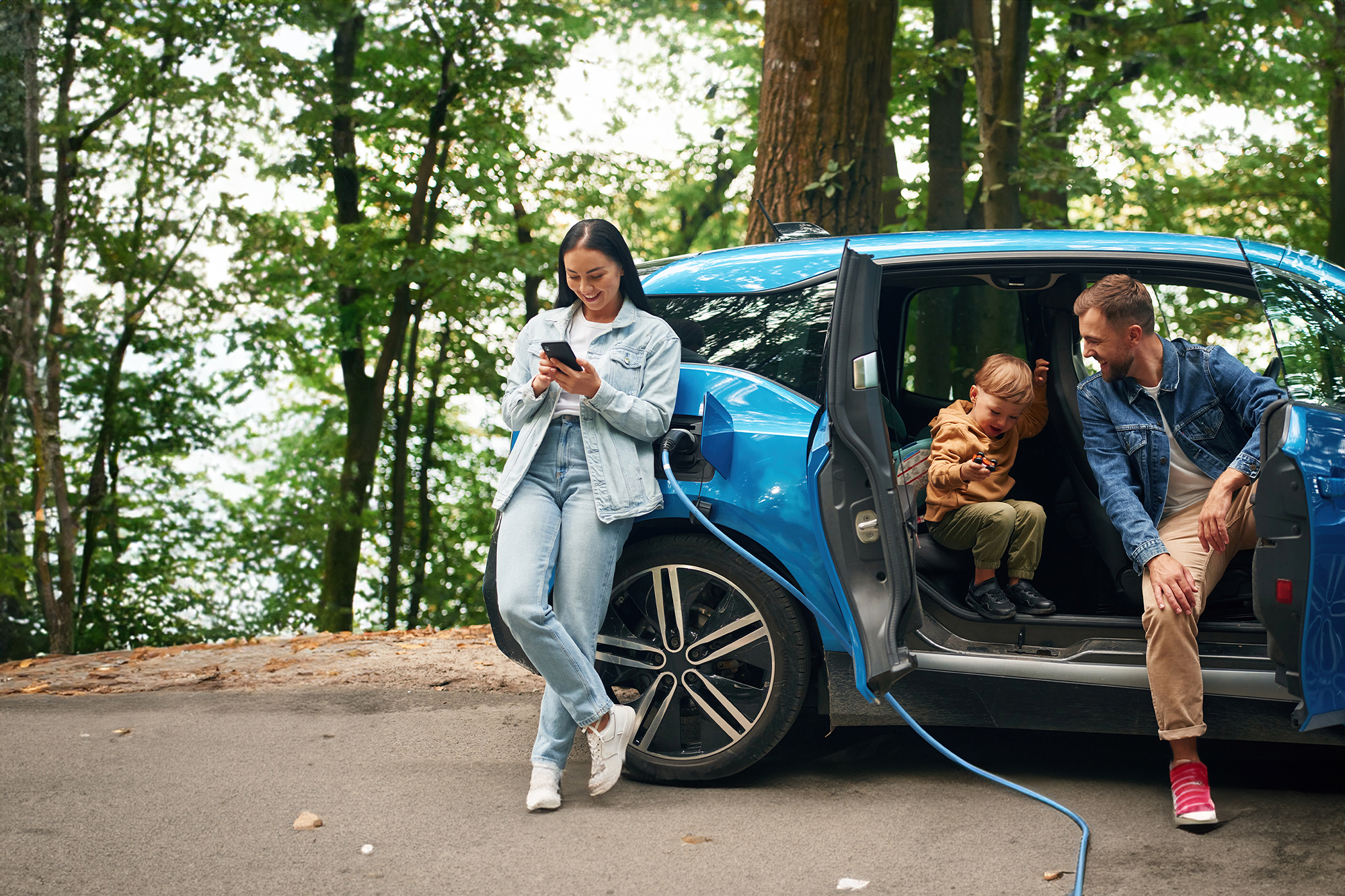 Een gezin bij een blauwe elektrische auto die wordt opgeladen, in een groene omgeving.